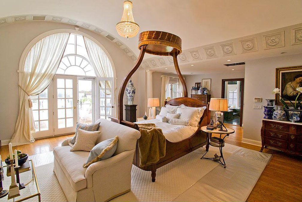 Elegant bedroom with a four-poster bed, sofa, arched windows, chandelier, and hardwood floors.