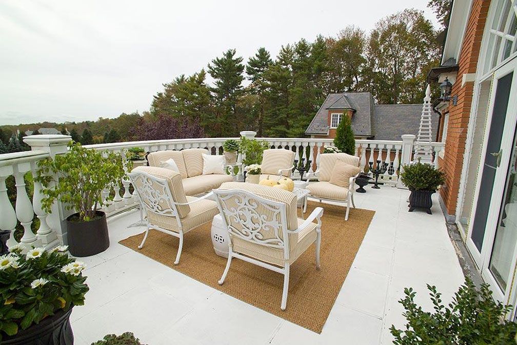 Outdoor patio with white furniture, rug, and potted plants on a white deck, overlooking trees.