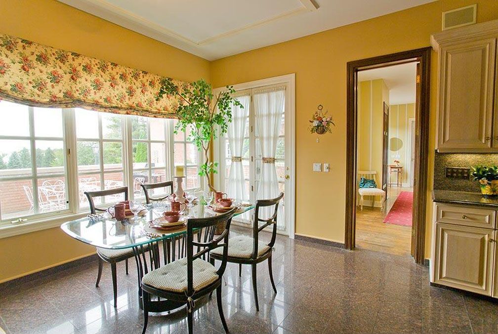 Dining area with a glass table, dark chairs, floral window covering, and access to the kitchen and another room.