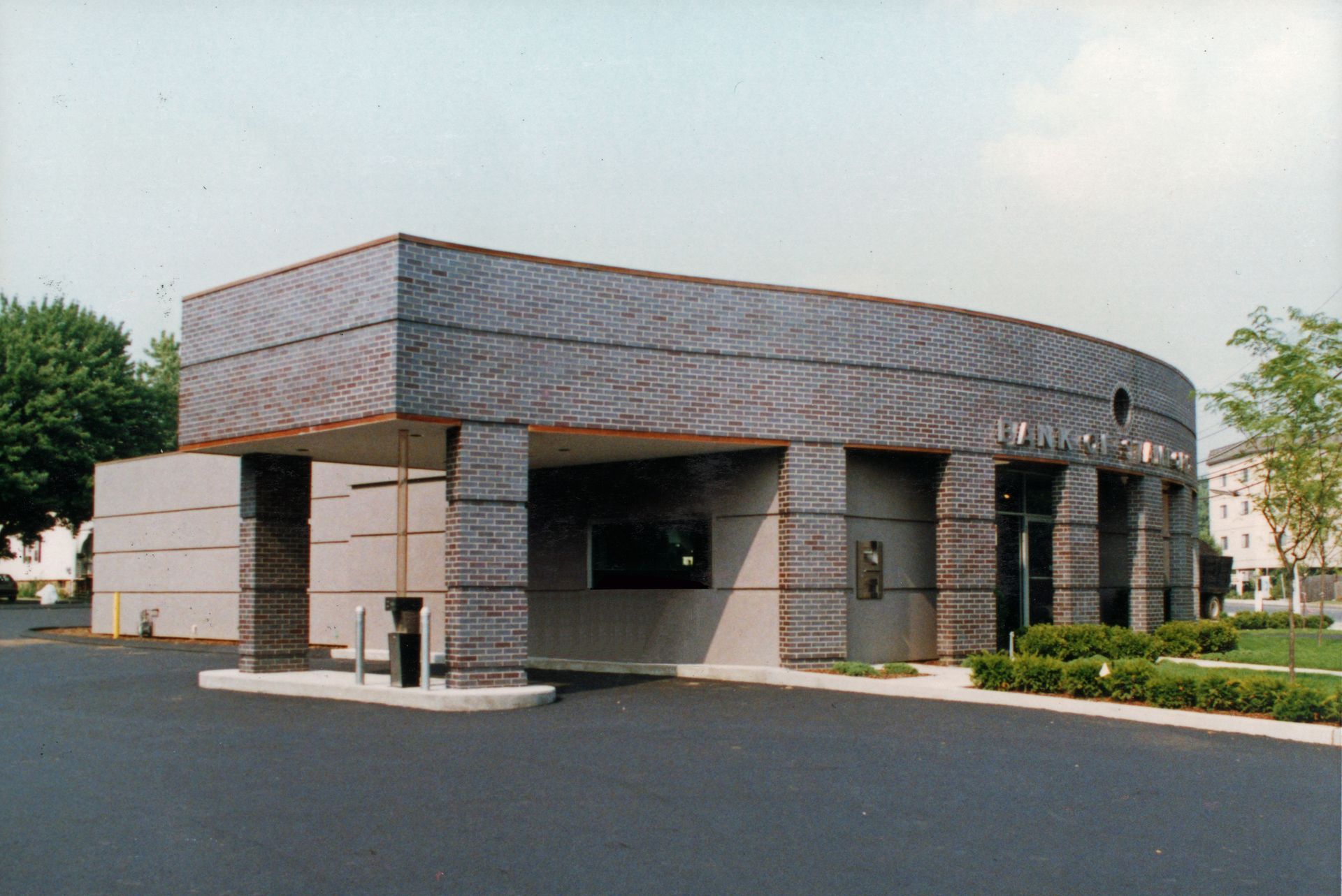 Bank building with curved roof, drive-through lane, dark asphalt, gray exterior, and landscaping.