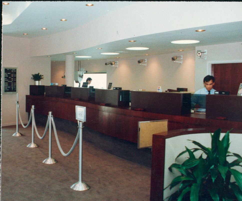 Bank interior with teller stations behind a long wooden counter, roped queue, and employees at work.