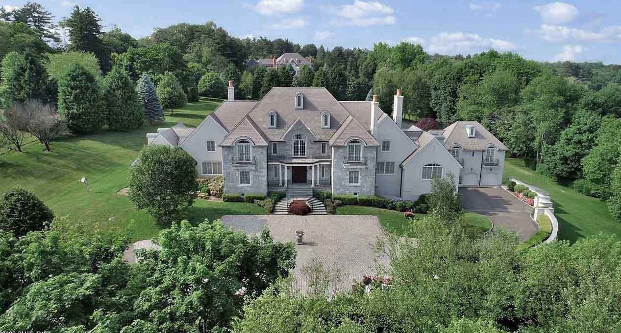 Large gray mansion with multiple gables and windows, set amongst green trees and lawn.
