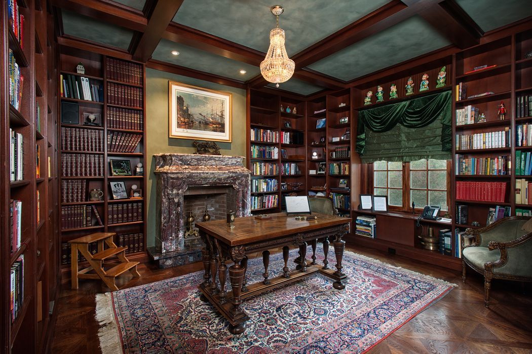 A wood-paneled library with a fireplace, ornate desk, bookshelves, rug, and chandelier.