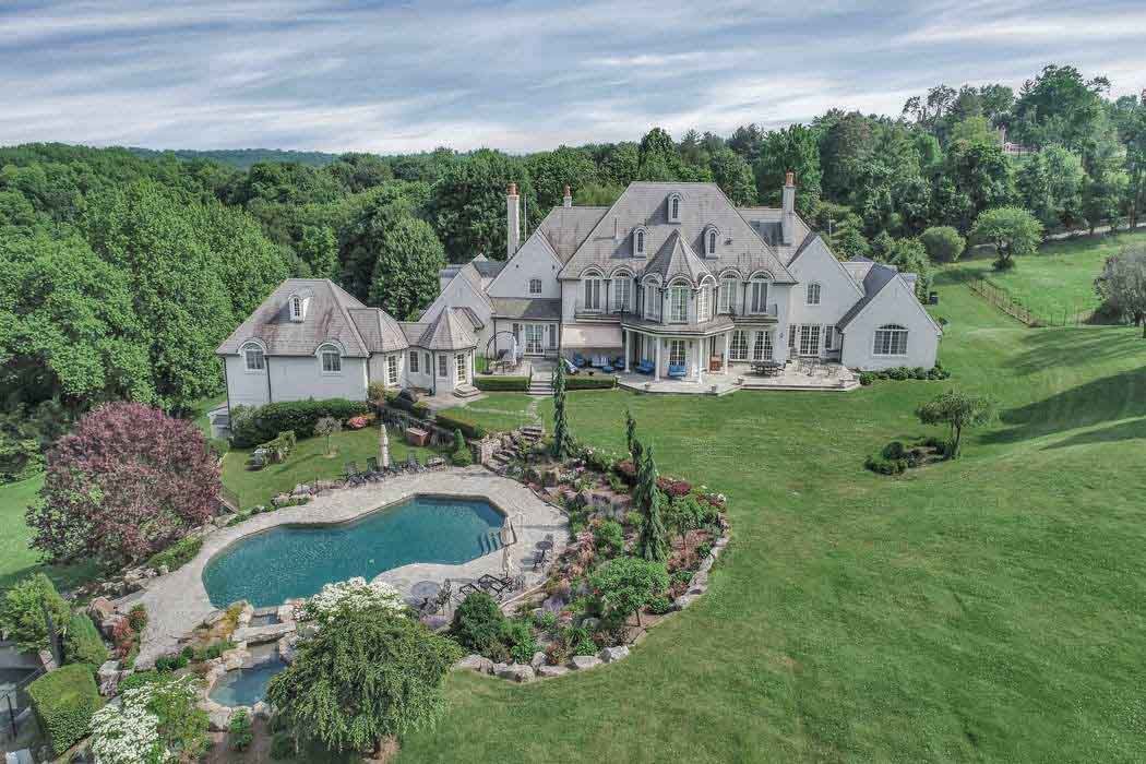 Large white house with a pool and surrounding greenery, nestled in a lush, green landscape.