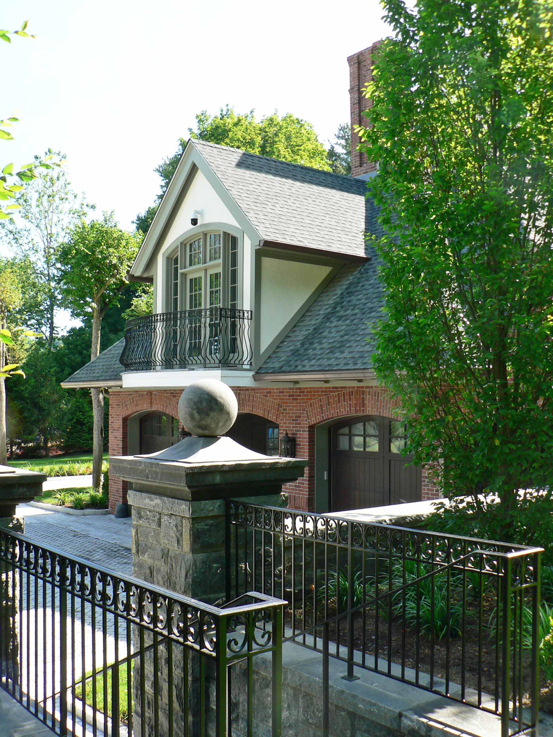 Brick garage with balcony and black wrought iron railing, set in a garden with trees.