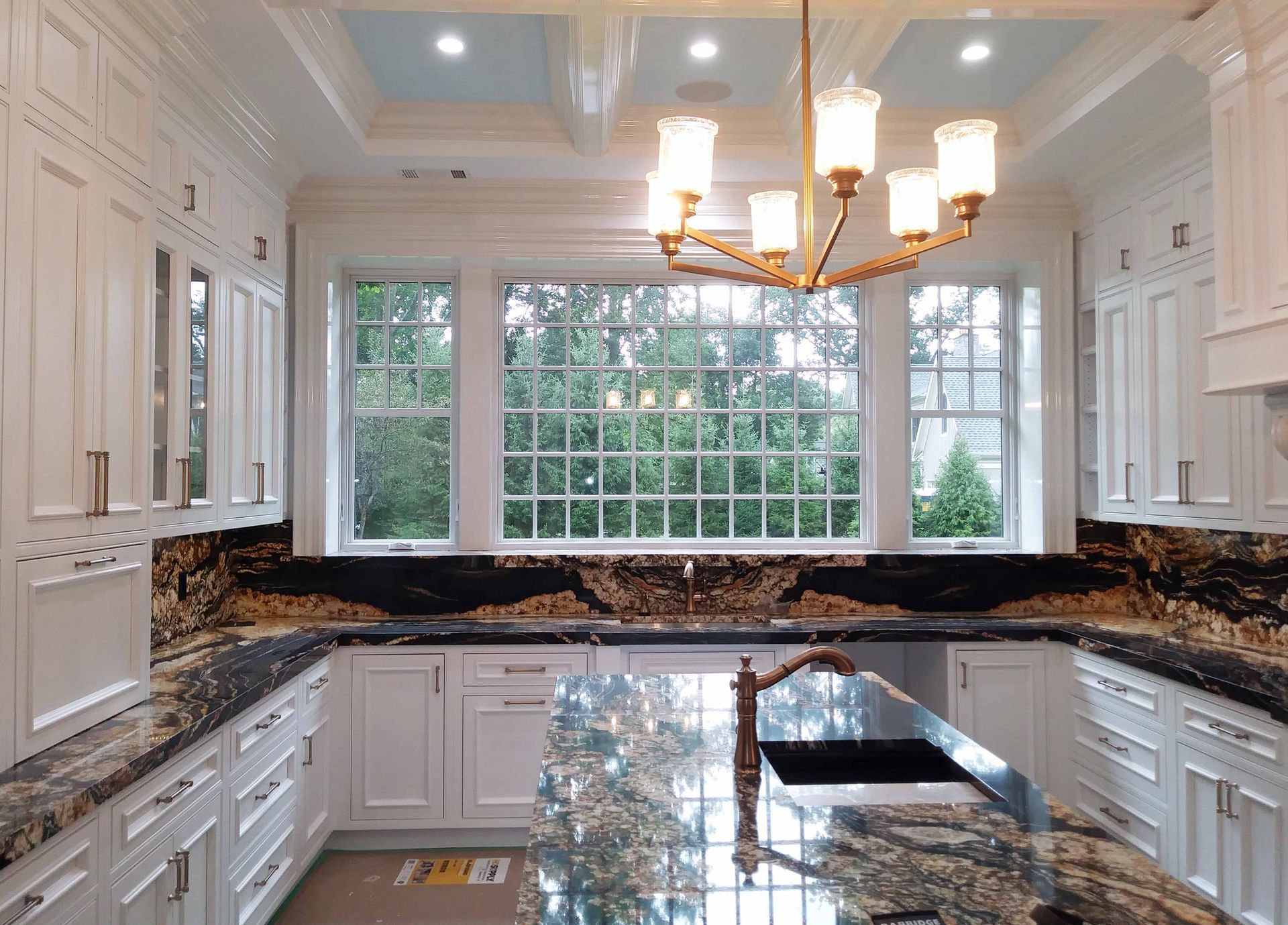 Elegant white kitchen with granite countertops, large windows, and a gold chandelier.