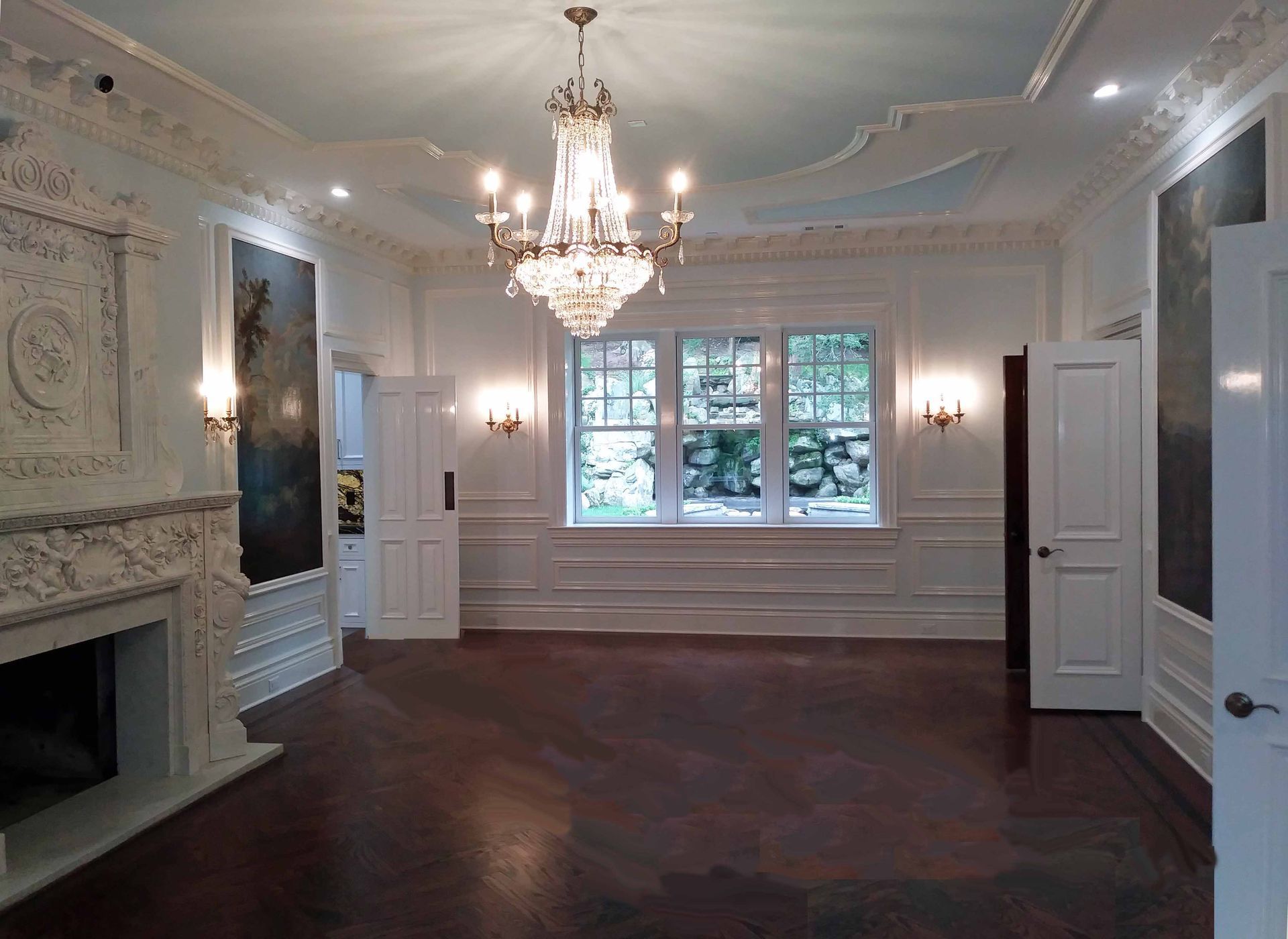 Ornate, empty room with chandelier, fireplace, windows, and dark floor. Light blue ceiling with decorative trim.