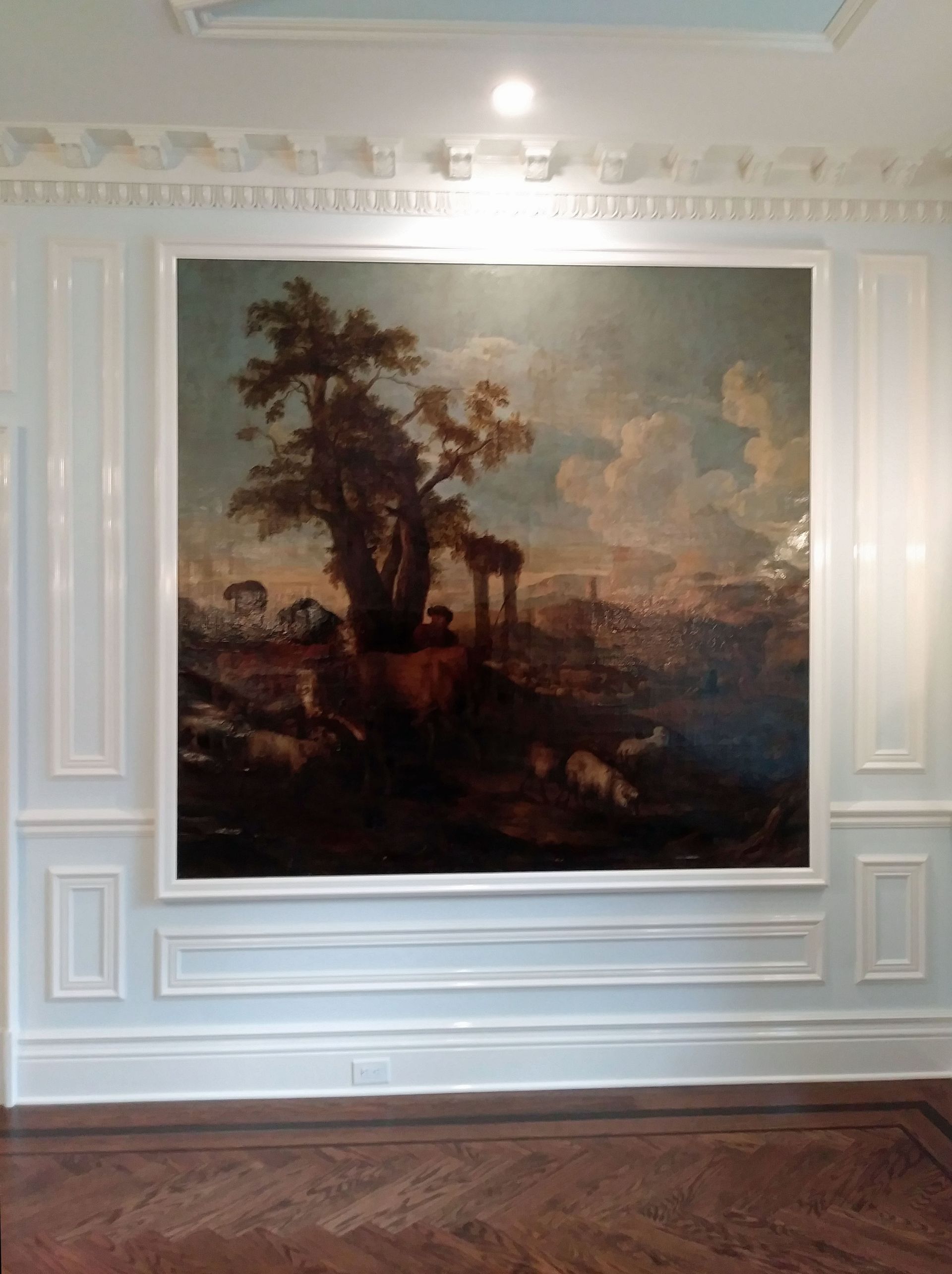 Large landscape painting with a tree, ruins, and cloudy sky, mounted on a white wall with molding.