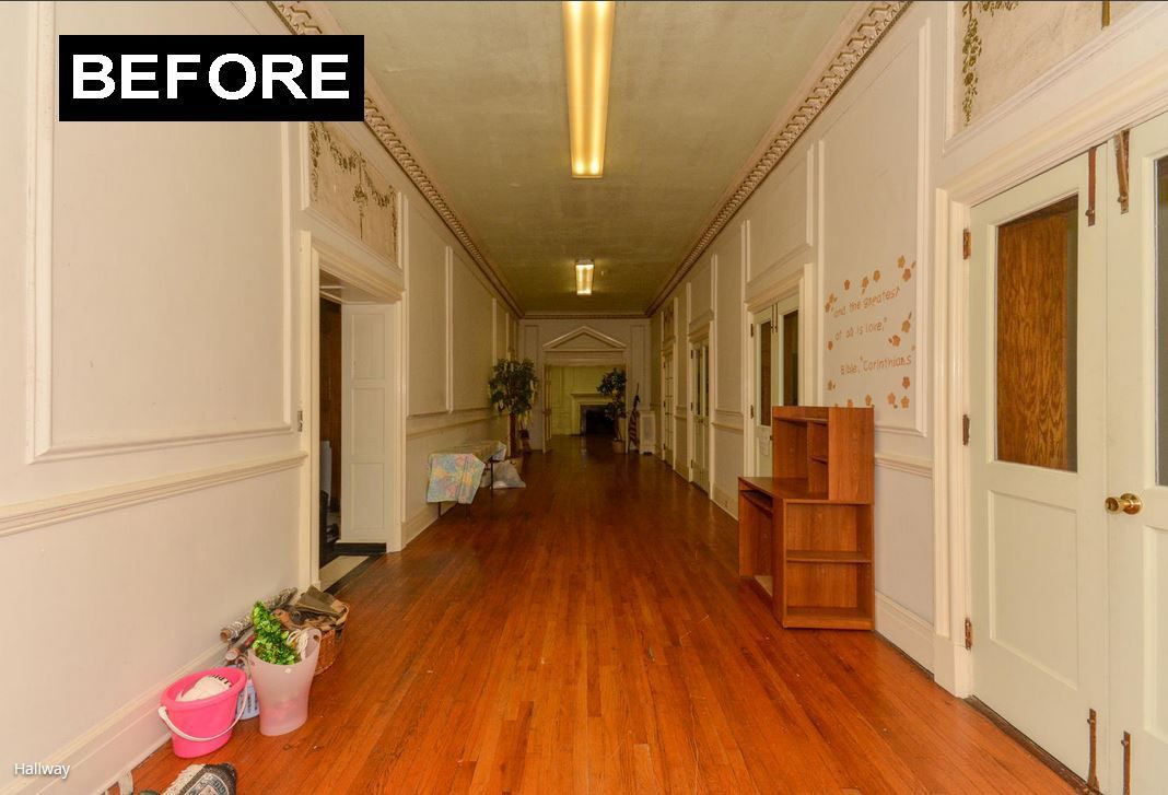 Long hallway with wooden floors, beige walls, and overhead lighting. Furniture and doors line the walls.