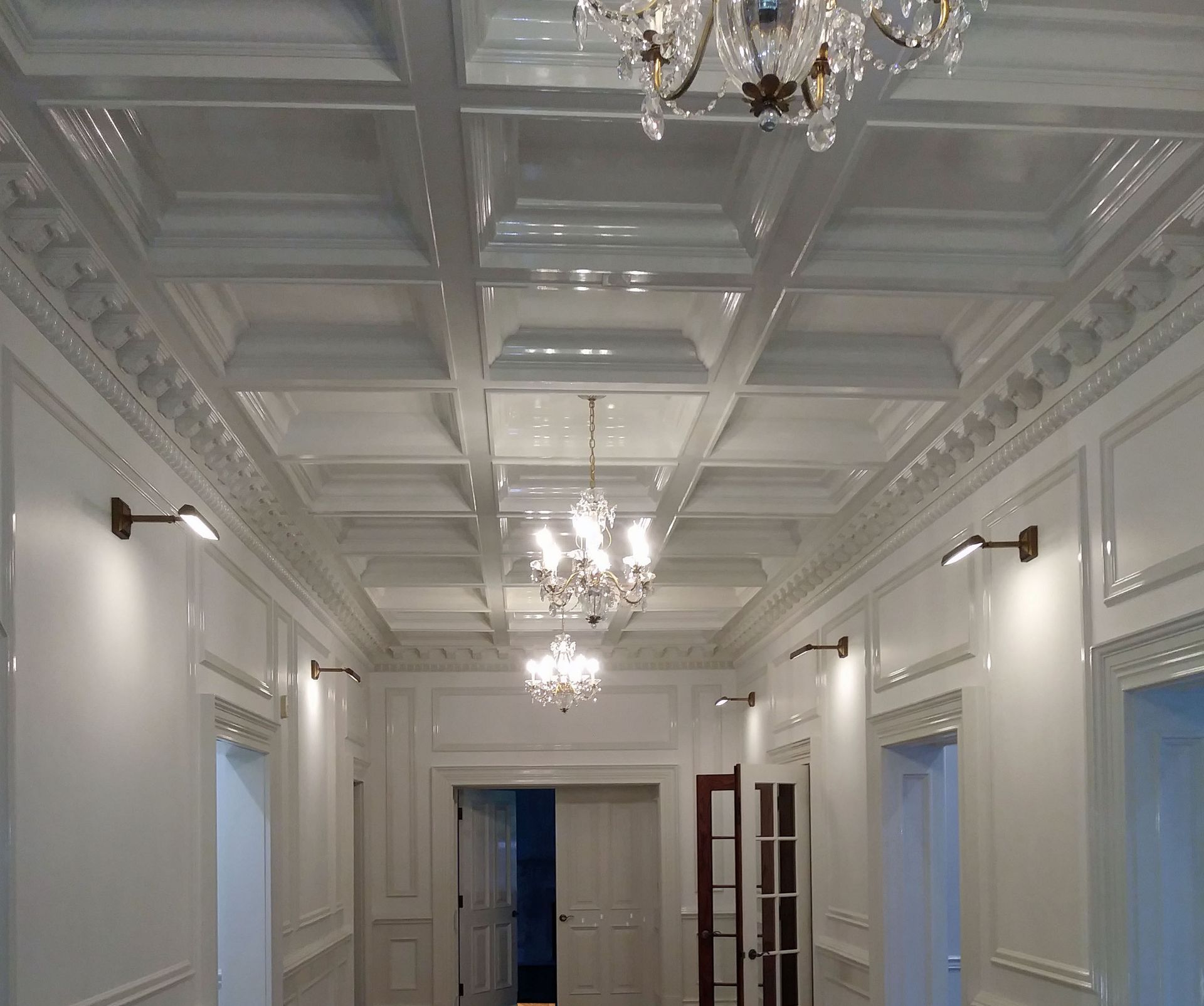 White hallway with coffered ceiling, chandeliers, and sconce lighting.