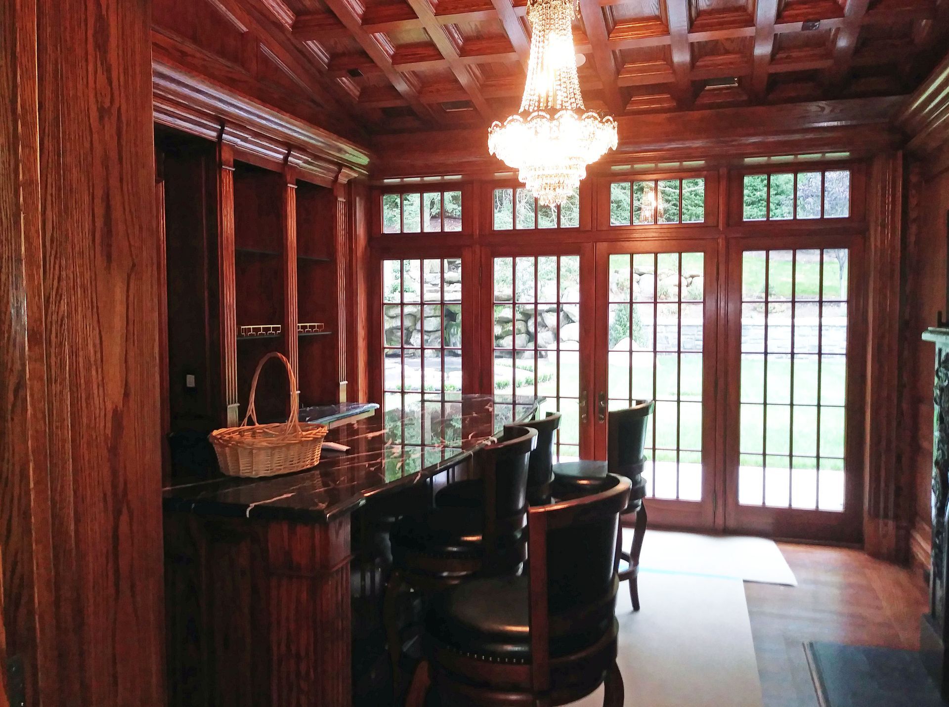 Dark wood bar with stools, built-in shelving, and a chandelier. French doors lead to a grassy outdoors.