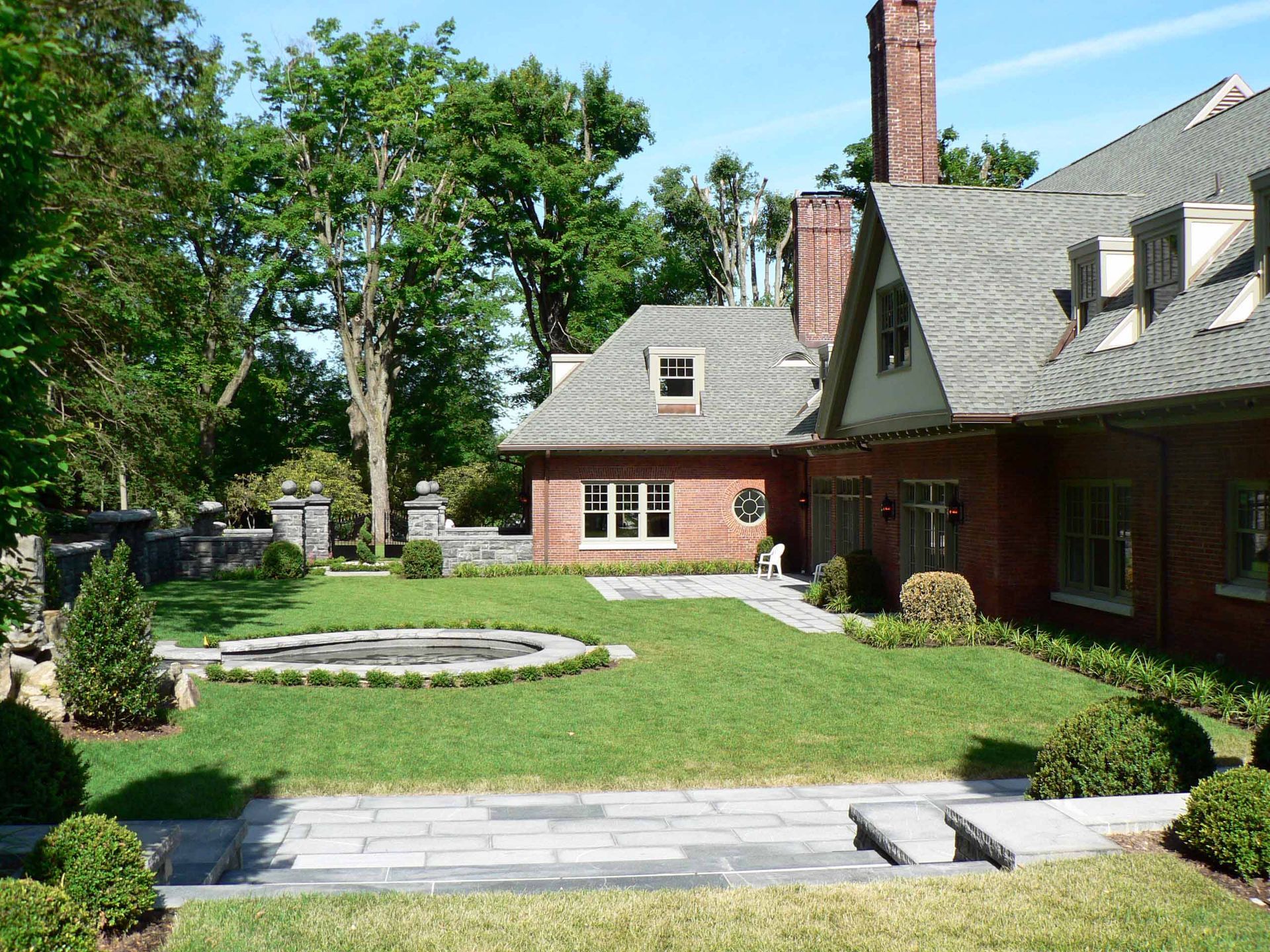Brick house with manicured lawn, stone patio, and round fountain in a sunny outdoor setting.