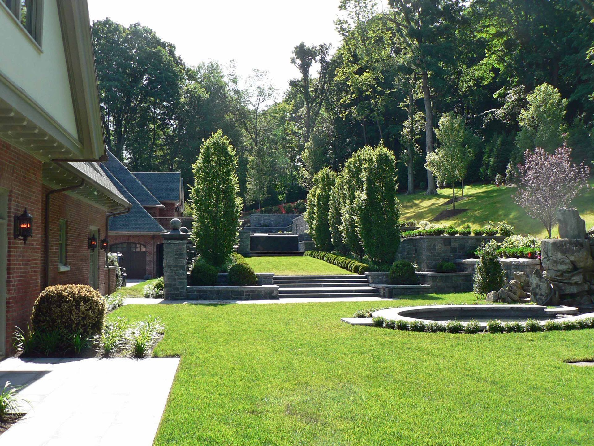 Green lawn and landscaped backyard with tiered stone walls, trees, and house.