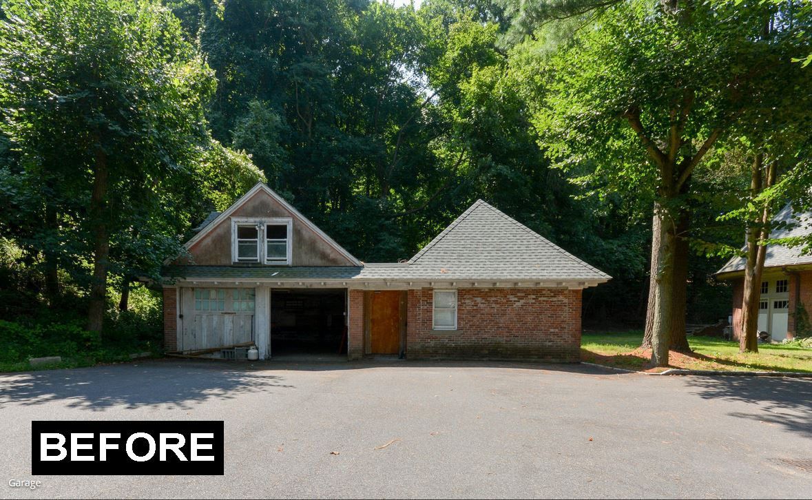 Old, weathered two-bay garage with gable roof and a red brick section; paved parking area and trees in background.