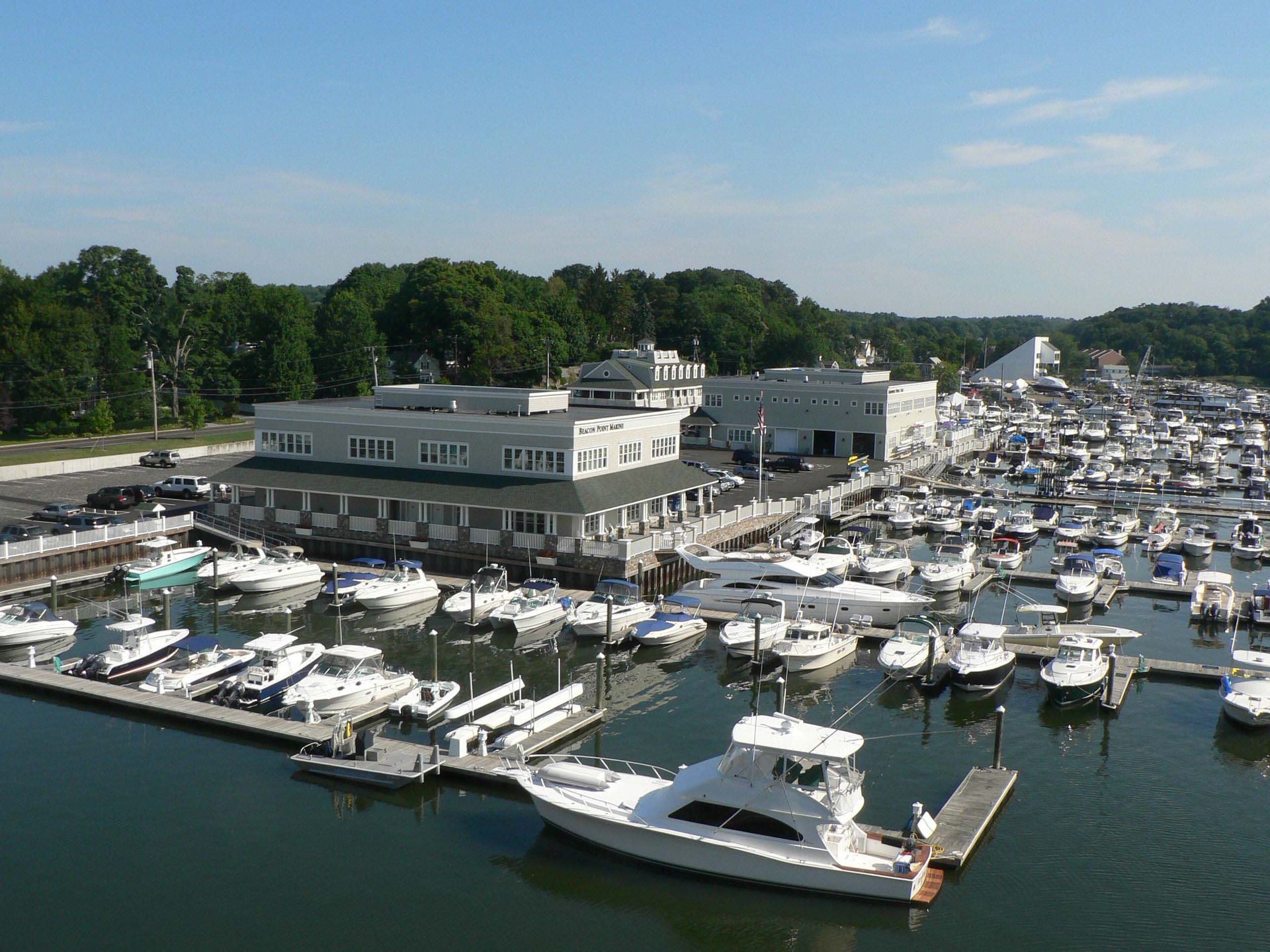 Boats docked at a marina with a multi-story building and trees in the background under a blue sky.