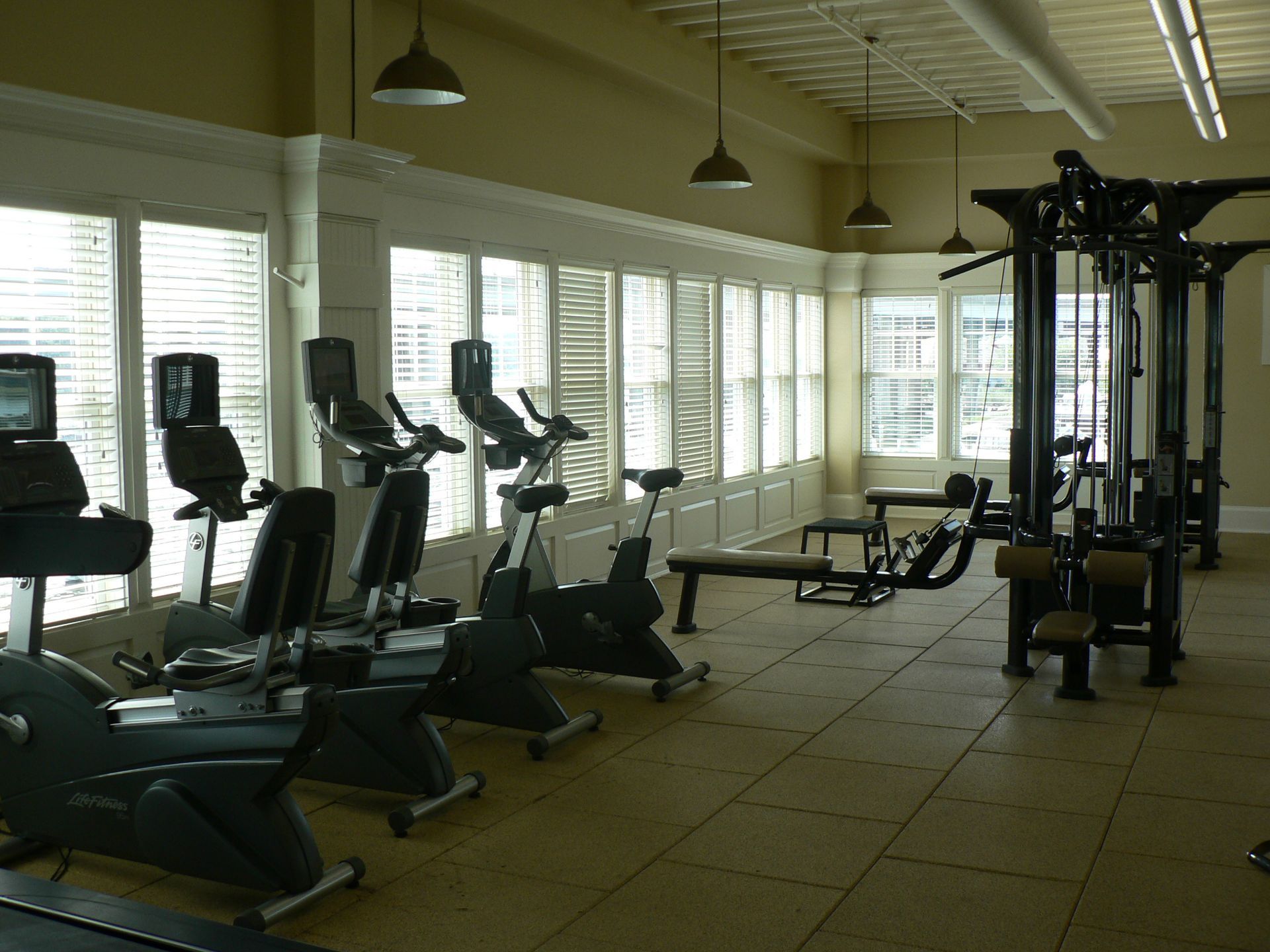 Gym interior with exercise bikes and weight machines near windows.