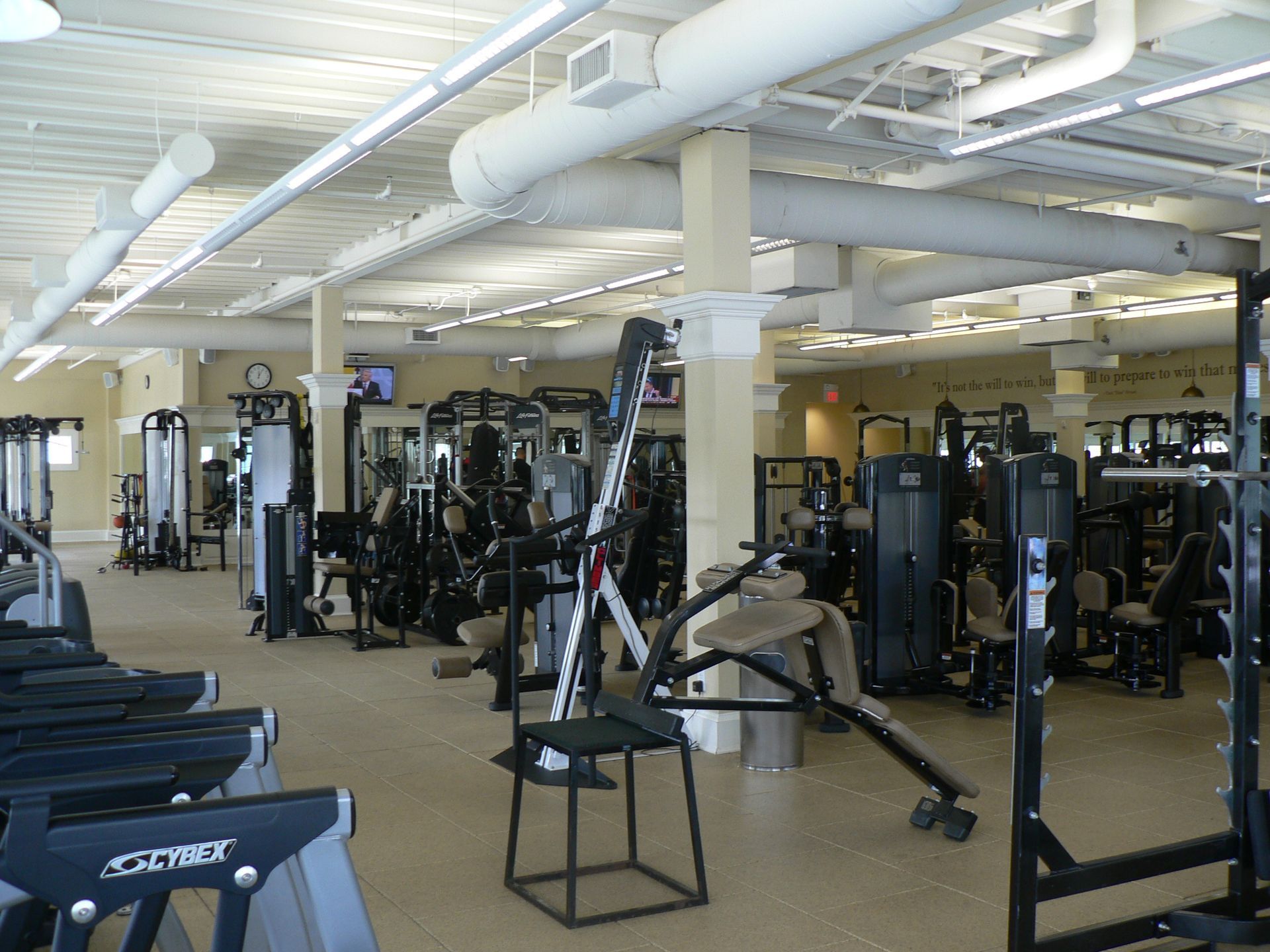 Gym interior with exercise machines and equipment under a white ceiling, overhead air ducts, and a tan floor.