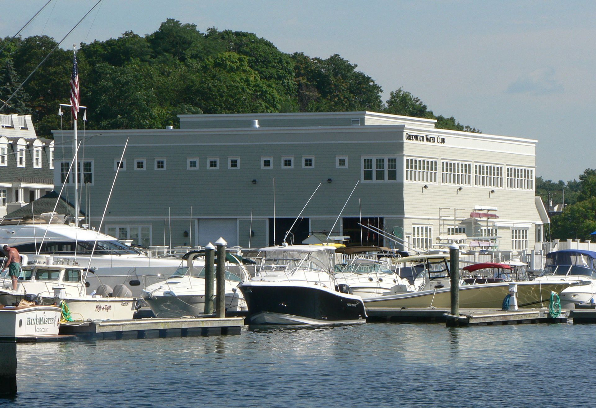 Boats docked at a marina with a light-colored building in the background. Green trees in the distance.