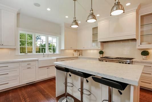 White kitchen with island, countertops, cabinets, and three bar stools. Hardwood floor and window.