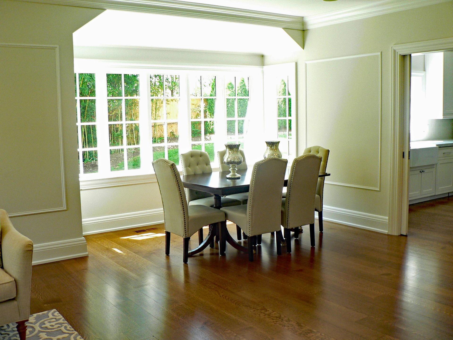 Dining room with dark wooden table, six beige chairs, and bay window overlooking greenery.