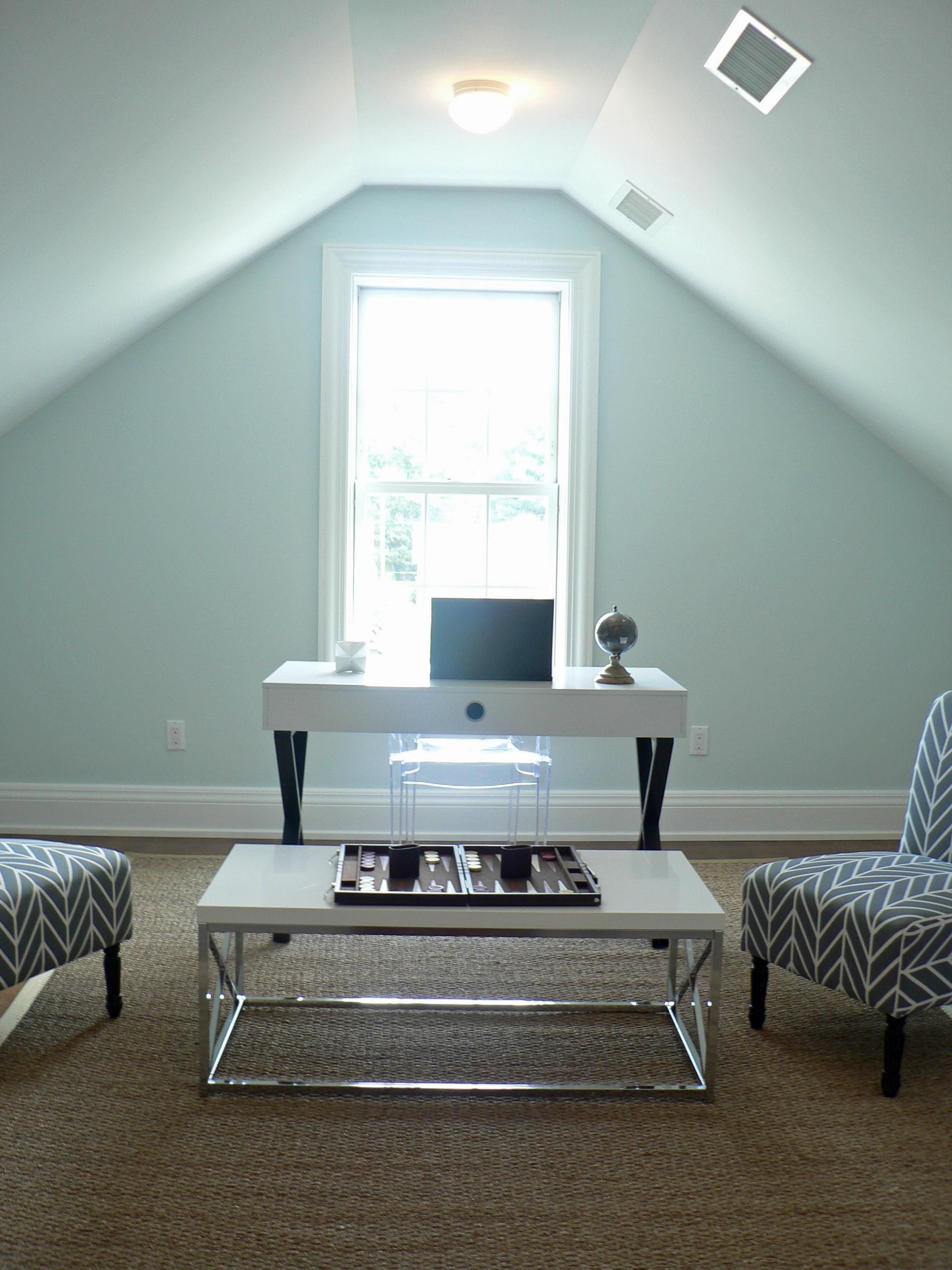 Attic office space with white desk, two patterned armchairs, and a coffee table. Window and light blue walls.