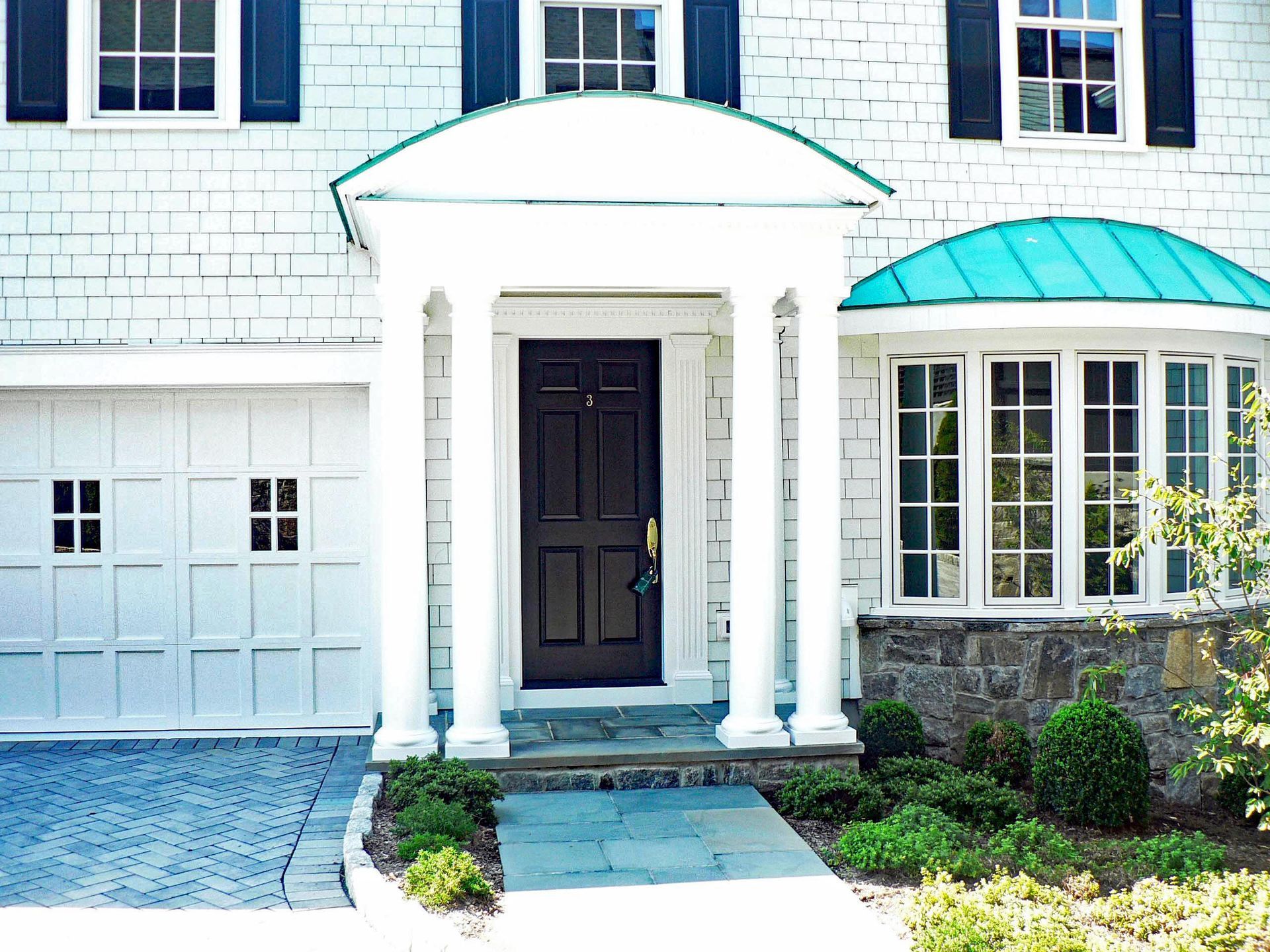White house with dark door, portico, and blue-green roof. Garage on the left, windows on the right.