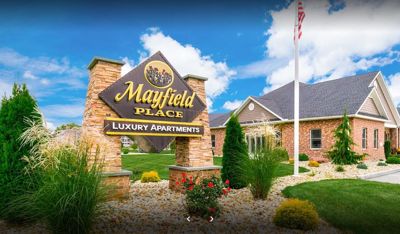 Sign for Mayfield Place Luxury Apartments with building and flag in background.