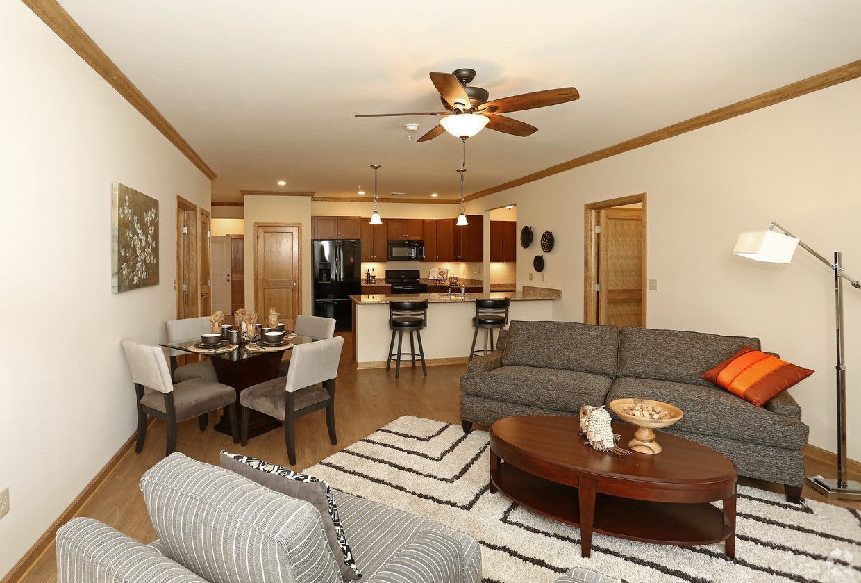 Living room with kitchen in the background; dining table, sofa, chairs, rug, and ceiling fan.