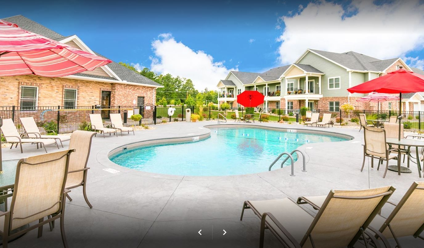 Pool with lounge chairs and umbrellas in front of apartment buildings on a sunny day.