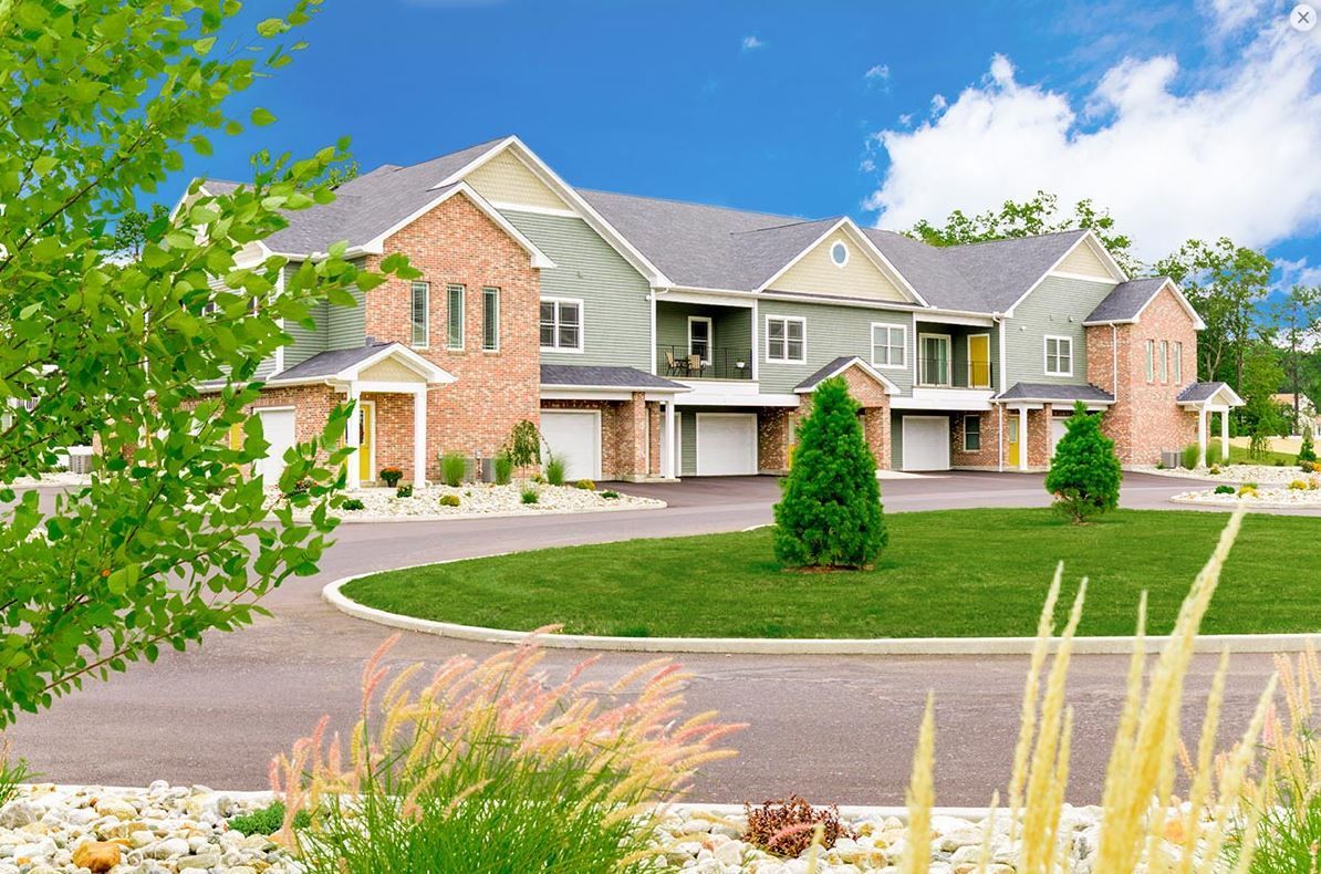 Townhouses with brick and green siding, driveway, and landscaping under a blue sky.