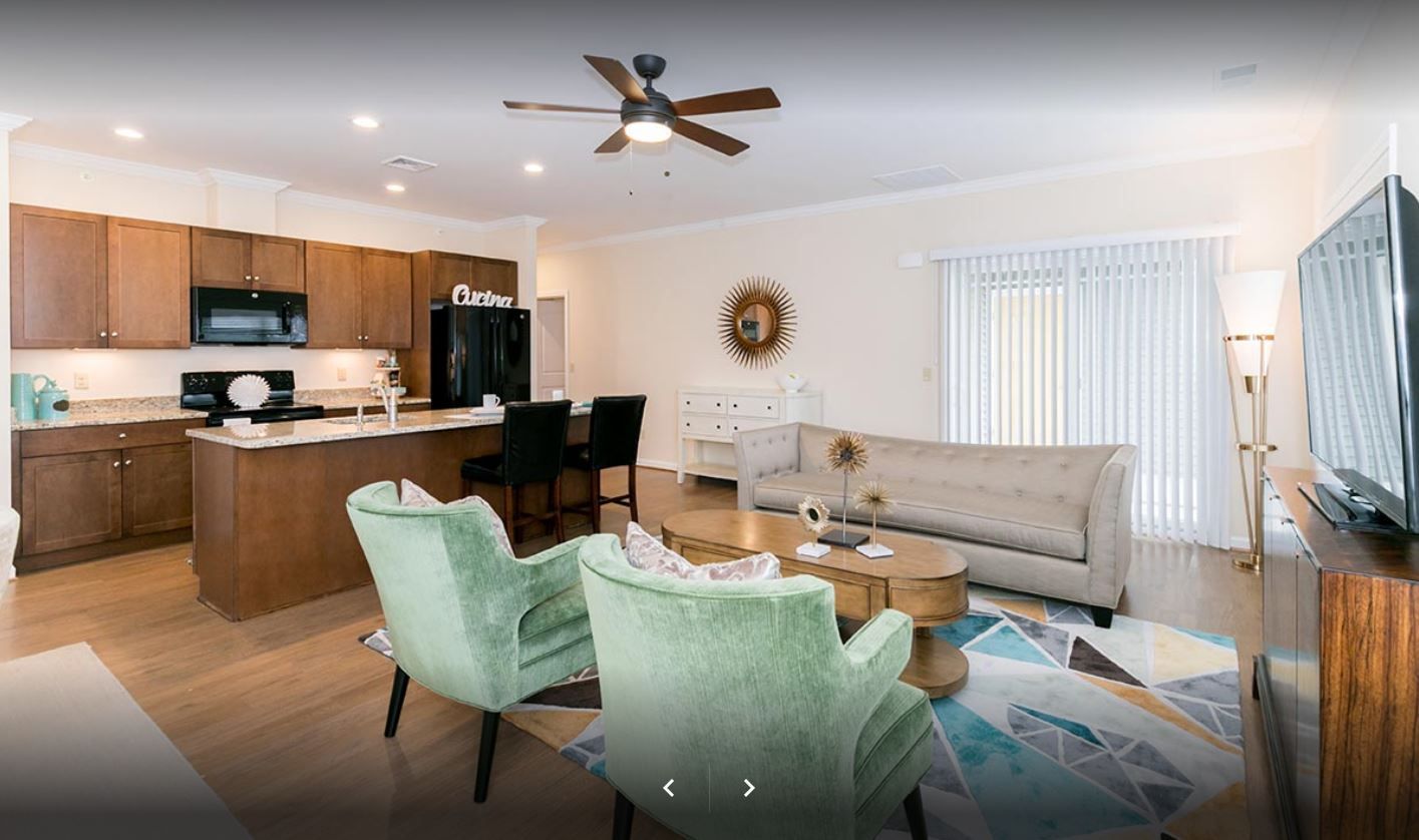 Living room with kitchen. Light wood cabinets, teal chairs, beige sofa, and a geometric rug.