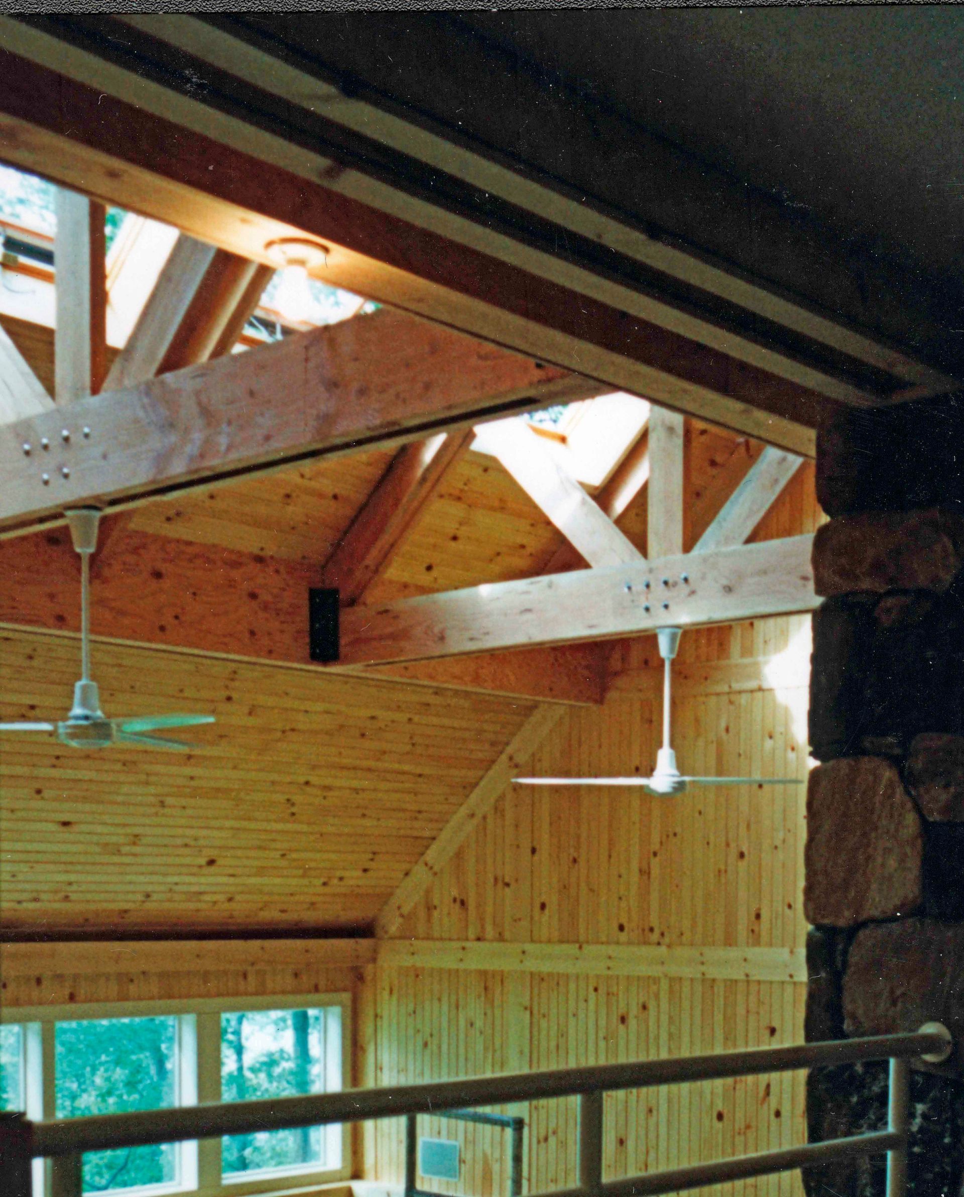 Interior view of a wood-paneled space with exposed beams, ceiling fans, and a stone column. Sunlight streams in.