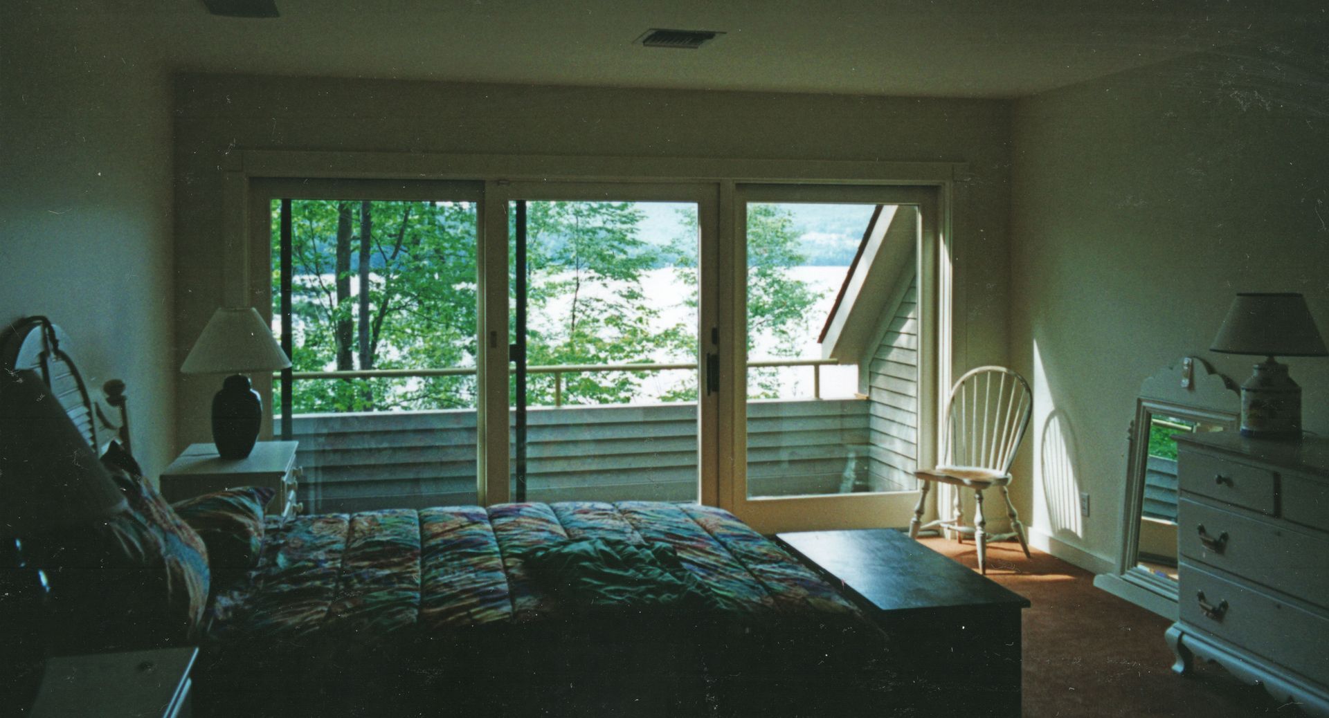 Bedroom with a bed, nightstands, and a chest of drawers. Sliding doors open to a balcony overlooking trees and water.