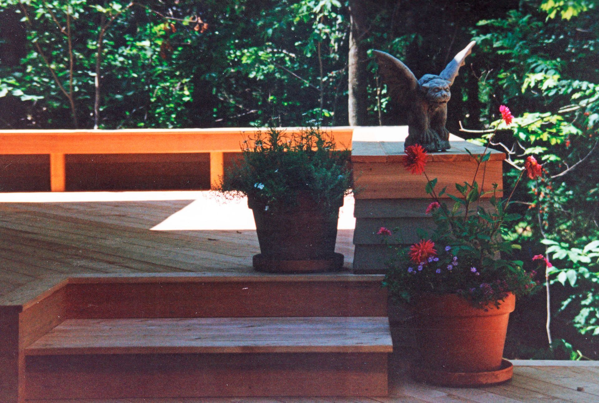 Wooden deck with steps, potted plants, and a gargoyle statue against a backdrop of trees.