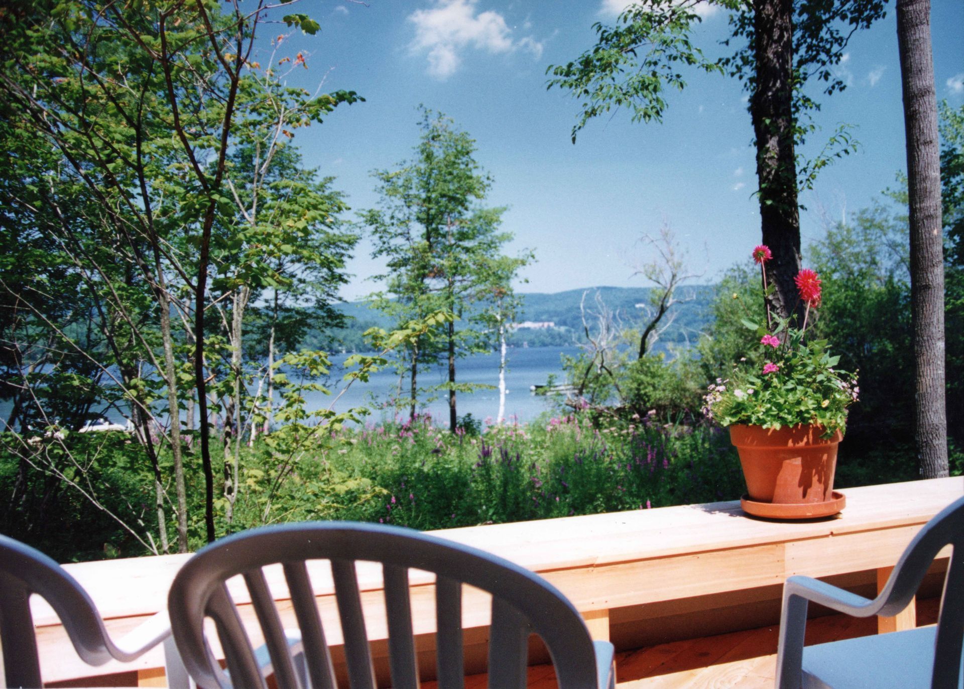 Patio overlooking a lake, with trees and a flower pot. Blue sky with clouds. White plastic chairs.