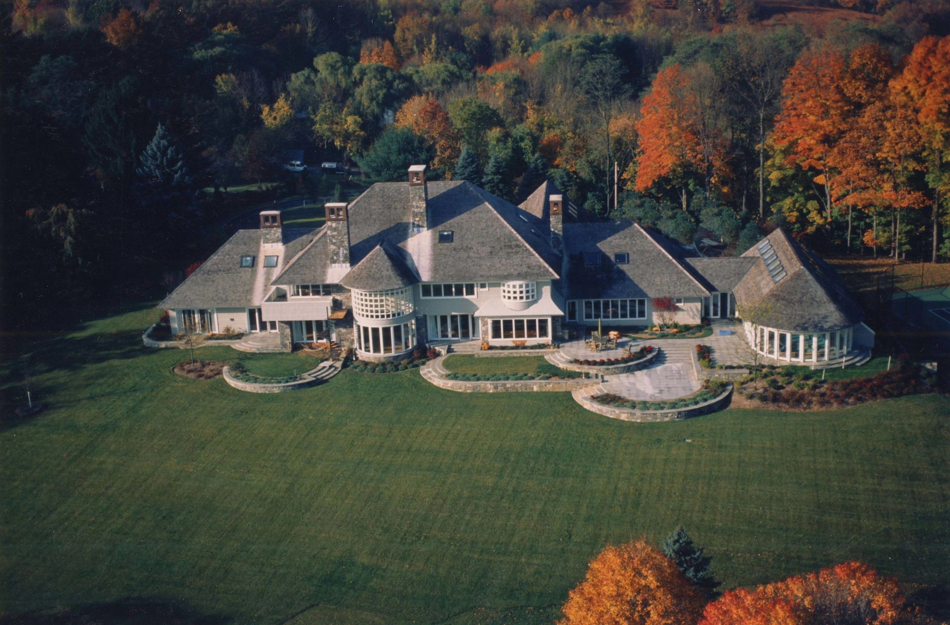 Large multi-winged house with gray roof on a green lawn, surrounded by fall foliage.