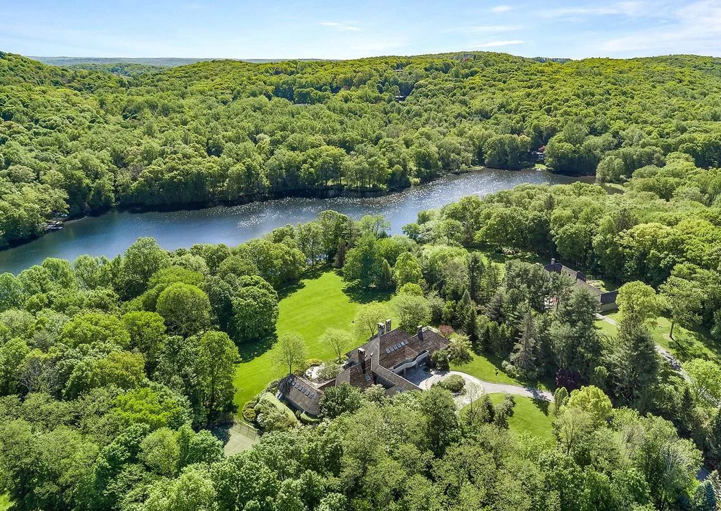 Aerial view of a house next to a lake surrounded by lush green trees. Sunny day.