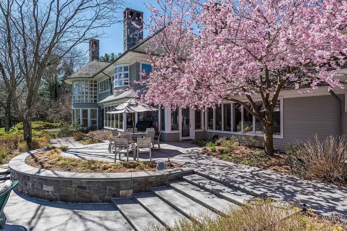 House with patio, stairs, and blossoming pink tree. Gray siding, stone patio and steps.
