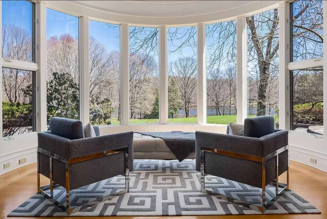 Two gray armchairs face a curved window overlooking a spring landscape; a patterned rug.