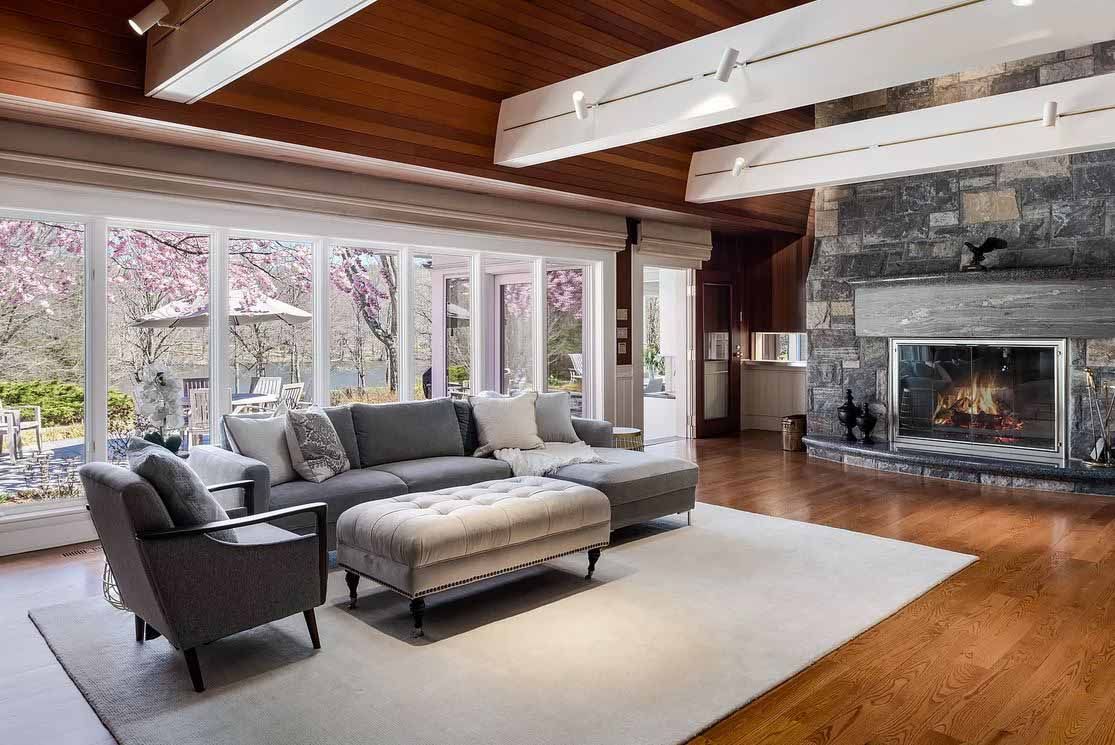 Living room with a large stone fireplace, gray sofa, and windows overlooking a garden.