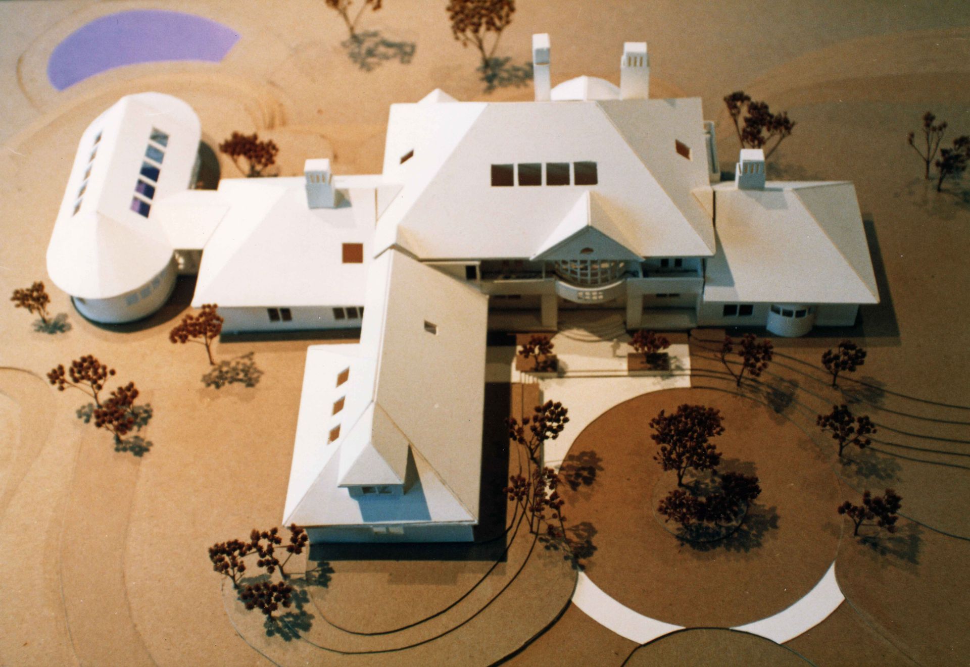 Architectural model of a white house with multiple wings and a circular pool, set on a beige surface with miniature trees.