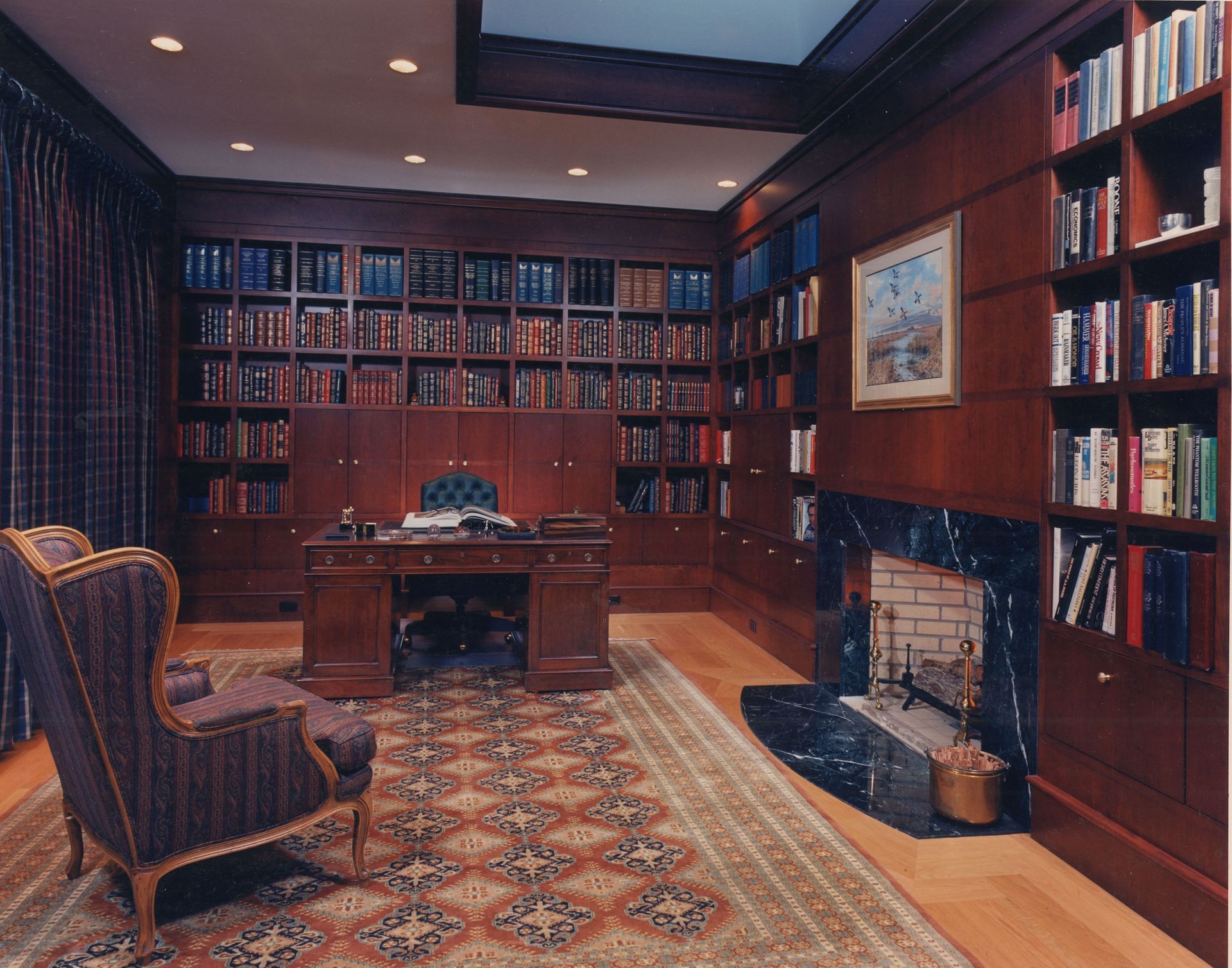 Library with dark wood paneling, built-in bookshelves, fireplace, desk, and patterned armchair.