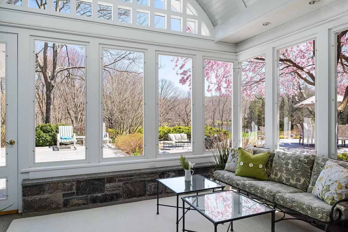 Sunroom with large windows overlooking a patio and trees with pink blossoms. Interior includes a couch and coffee tables.