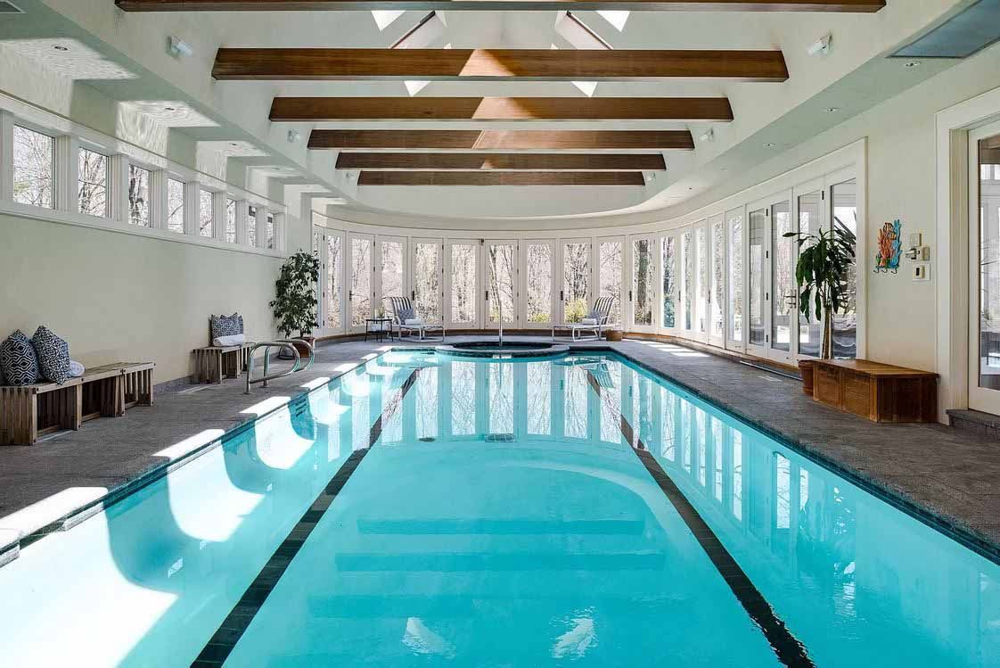 Indoor pool with arched windows, skylights, and wooden beams. Clear water and plants line the walls.
