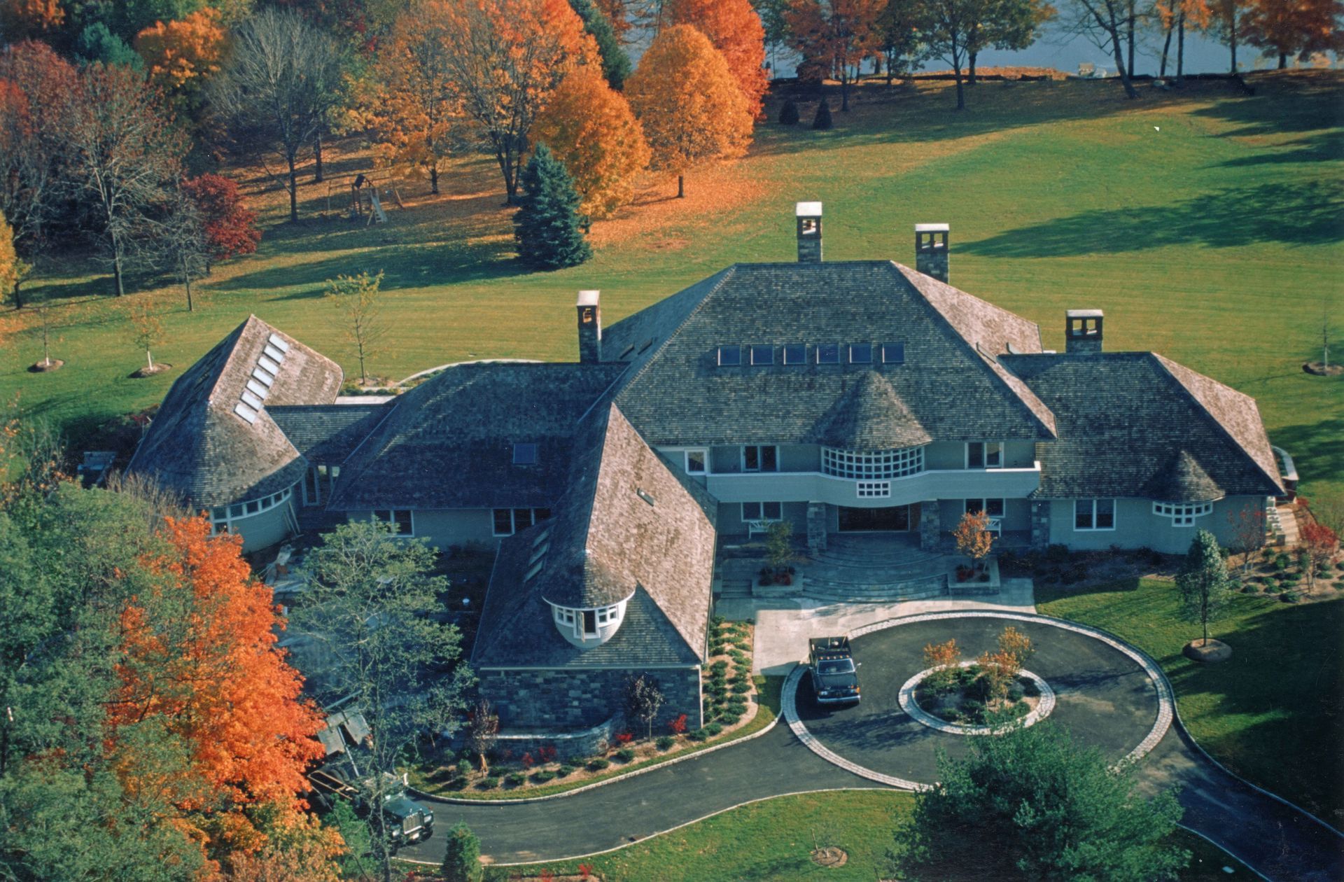 Large house with a shingled roof and stone facade set in fall foliage. A circular driveway leads to the entrance.