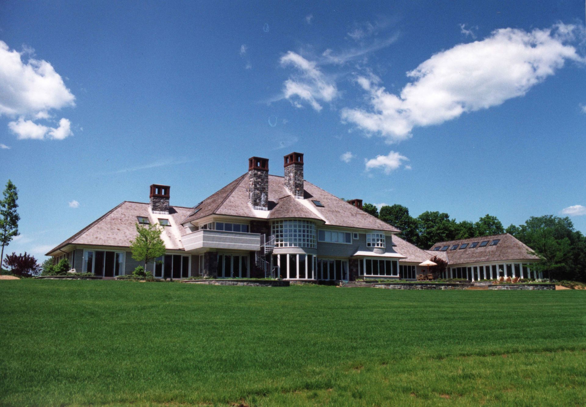 Large, light-colored house with a shingled roof, set on a green lawn against a blue sky with clouds.