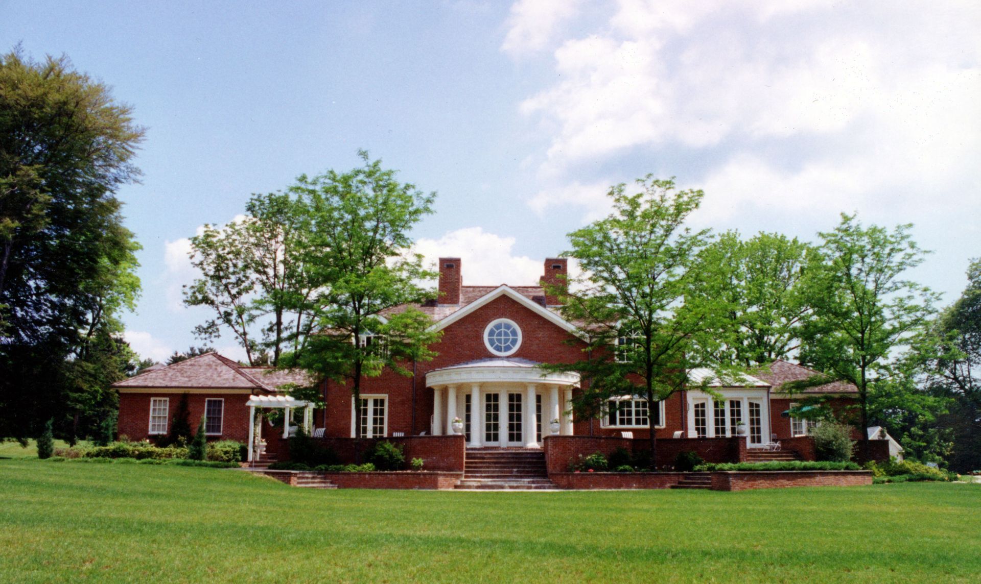 Red brick house with white trim, arched portico, and green lawn under a sunny sky.