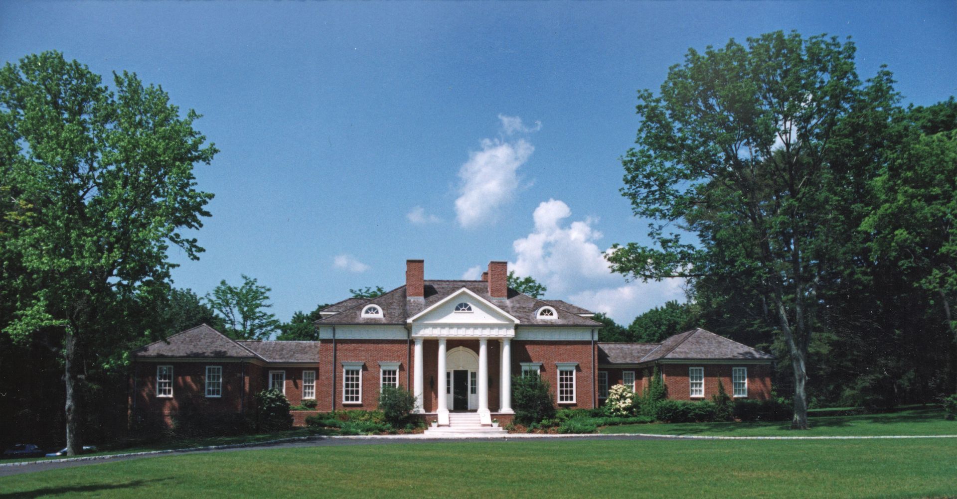 Red brick building with white columns and a central doorway, framed by trees on a sunny day.