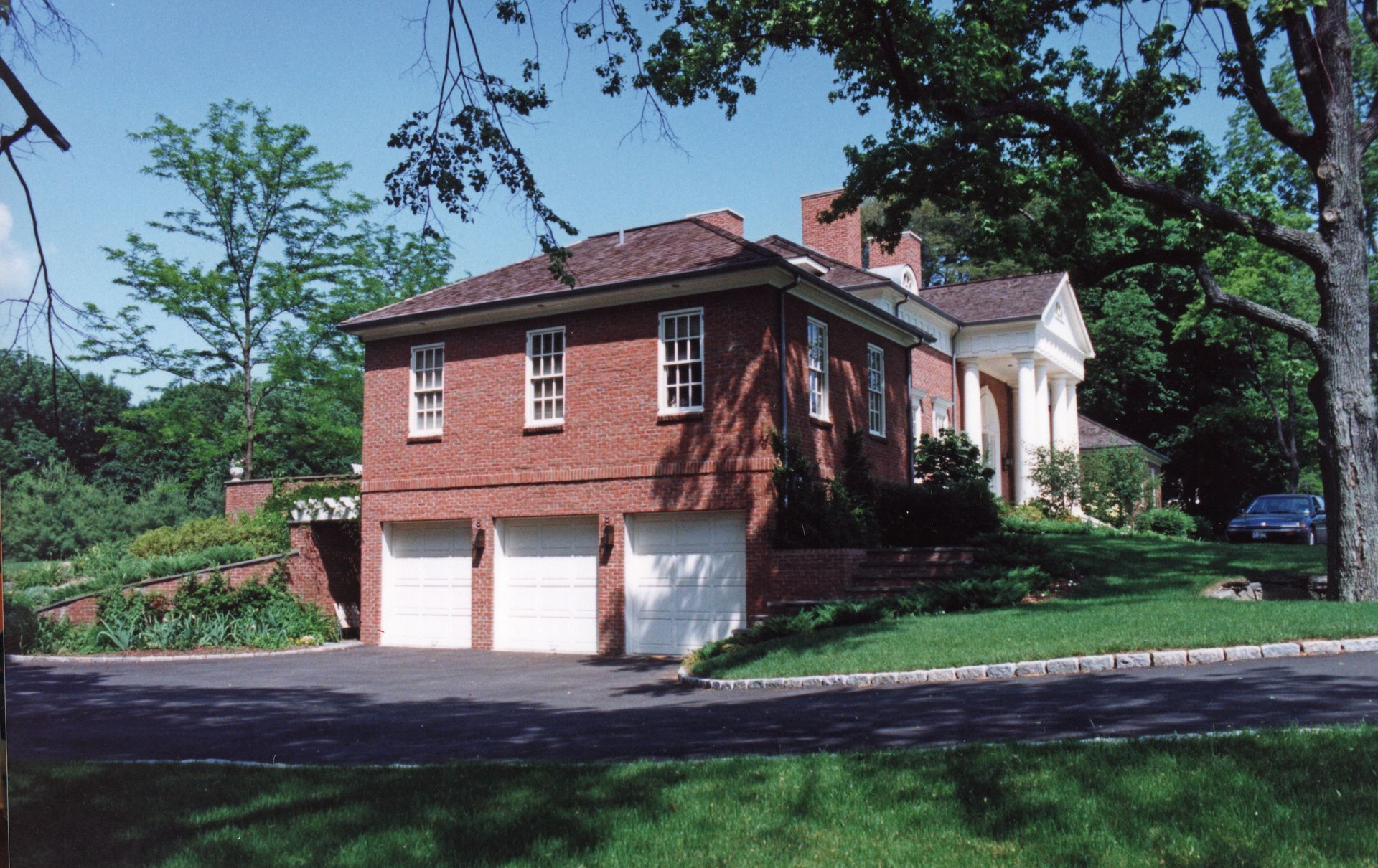 Red brick house with garage, white doors, and a classical portico. Green grass and trees surround it.