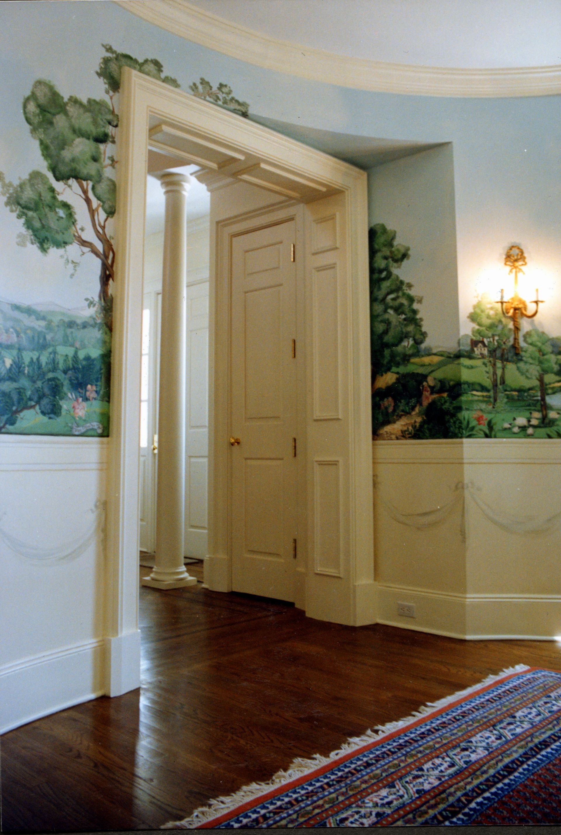 Interior with painted landscape on walls, doorway with columns, and wooden floors.